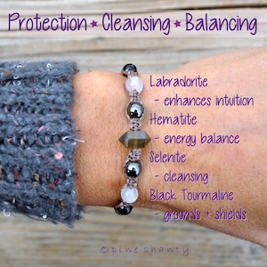 Puede incluir: Pulsera artesanal con cuentas de labradorita, hematita, selenita y turmalina negra. La pulsera se muestra en una muñeca. El texto de la imagen dice "Protección, Limpieza, Equilibrio".