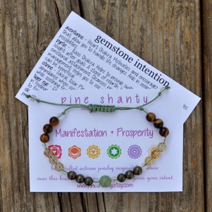May include: Green and brown gemstone bracelet with a card that reads "gemstone intention pine shanty Manifestation + Prosperity". The card also has a description of the stones and their meanings. The bracelet is handmade and features a variety of stones, including aventurine, pyrite, citrine, and tiger's eye.