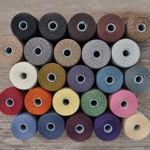 May include: A collection of 24 spools of thread in various colors, including beige, brown, gray, blue, green, red, orange, yellow, and purple. The spools are arranged in a semi-circle pattern on a wooden surface.
