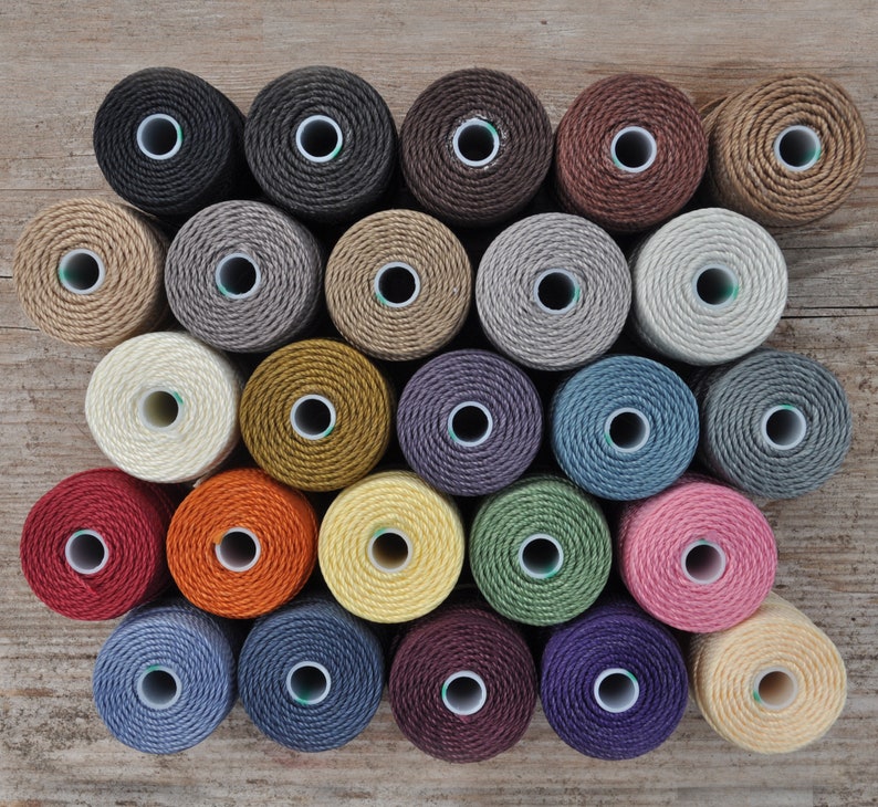 May include: A collection of 24 spools of thread in various colors, including beige, brown, gray, blue, green, red, orange, yellow, and purple. The spools are arranged in a semi-circle pattern on a wooden surface.