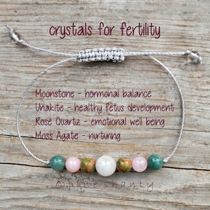 May include: A gray string bracelet with seven beads, including moonstone, unakite, rose quartz, and moss agate. The bracelet is on a wooden surface with the text "crystals for fertility" above the bracelet and the text "Moonstone - hormonal balance", "Unakite - healthy fetus development", "Rose Quartz - emotional well being", "Moss Agate - nurturing", and "Opine shanty" below the bracelet.
