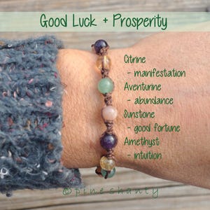 May include: A brown leather bracelet with seven gemstone beads. The beads are citrine, aventurine, sunstone, and amethyst. The bracelet is tied with a knot and has the text "Good Luck + Prosperity" at the top and "@pine shanty" at the bottom.