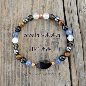May include: A bracelet made of various coloured stone beads, including black, brown, white, and blue. The bracelet features a black, oblong stone and copper-coloured spacers. Text on the image reads "empath protection + EMF shield".