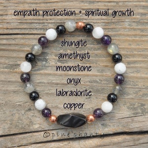 May include: A bracelet made with black onyx, labradorite, amethyst, moonstone, shungite, and copper beads. The bracelet is on a wooden surface and has the text "empath protection + spiritual growth" and "@pineshanty" on it.