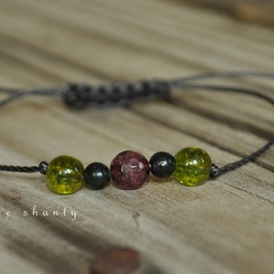 May include: A black cord bracelet with green and red beads. The bracelet has a sliding knot for adjusting the size.