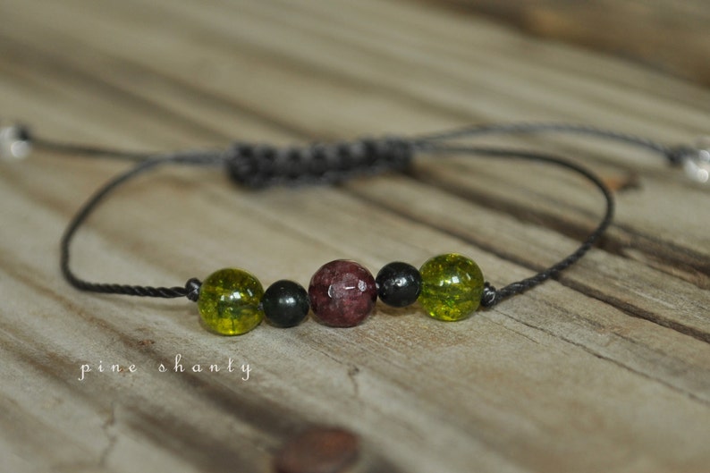 May include: A black cord bracelet with green and red beads. The bracelet has a sliding knot for adjusting the size.