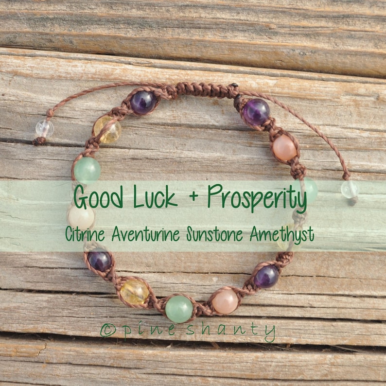 May include: A brown string bracelet with citrine, aventurine, sunstone, and amethyst beads. The bracelet is on a wooden surface and has the text "Good Luck + Prosperity Citrine Aventurine Sunstone Amethyst" on a green background.