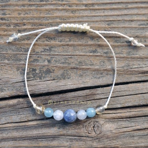 May include: A white string bracelet with blue and clear gemstone beads. The bracelet has a sliding knot closure.