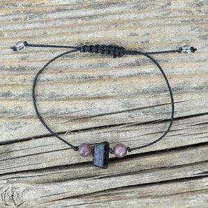 May include: Black bracelet with a black tourmaline stone and two pink beads. The bracelet is made with black string and has clear beads on the ends.