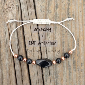 May include: A white string bracelet with black and copper beads and a black obsidian stone. The bracelet is labeled "grounding + EMF protection" and "pine shanty".