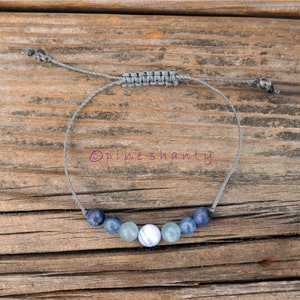 Puede incluir: Un brazalete de cuerda gris con cuentas de piedras preciosas azules y azul claro. El brazalete tiene un cierre de nudo deslizante. © Pineshanty