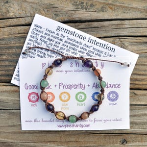 May include: A brown string bracelet with seven different colored gemstone beads. The bracelet is on a white card with the text "Pineshanty" and "Good Luck + Prosperity + Abundance".