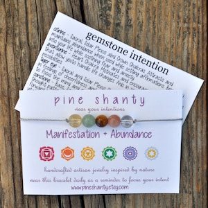 May include: A gray string bracelet with seven gemstone beads, including clear, green, brown, and pink. The bracelet is on a white card with the text "pine shanty wear your intentions" and "Manifestation + Abundance". The card also features seven colorful symbols representing the seven chakras.