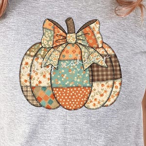 May include: A gray t-shirt featuring a patchwork pumpkin design with a bow. The pumpkin is composed of various fabric patterns in orange, green, and brown. The text "digital download no physical product included" is printed on the shirt.