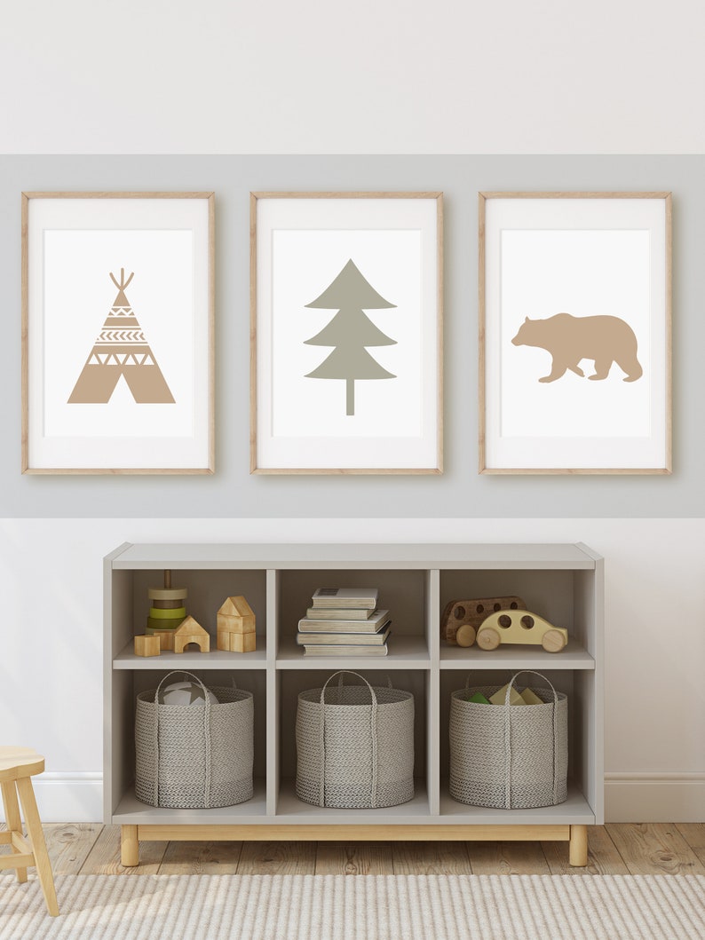 May include: Three framed prints on a light gray wall. The prints feature a brown teepee, a green tree, and a brown bear. A gray wooden shelf with six cubbies is below the prints. The cubbies contain wooden toys, books, and gray woven baskets.