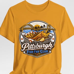 Könnte beinhalten: Senfgelbes T-Shirt mit einem kreisförmigen Aufdruck eines Fischbratens. Die Grafik zeigt einen Fisch, Pommes Frites und eine Zitronenscheibe in einem Korb, mit Brücken und dem Text "Pittsburgh Fish Fry Club".
