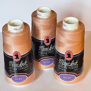 以下が含まれることがあります： ピーチ色のMaxi-Lockロックミシン糸3本。各スプールには、Maxi-Lock Serger Thread、3000 YDS、2742 Mと書かれた白い文字の黒いラベル、ピンクのミシンのアイコン、紫色の値札が付いています。