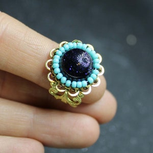 May include: A gold ring with a blue gemstone surrounded by turquoise beads. The ring is on a finger.