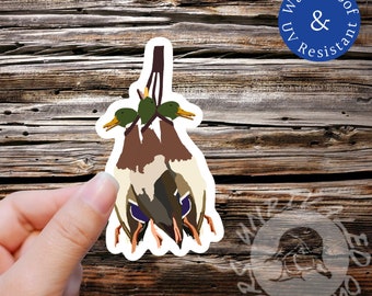 May include: A sticker of three mallard ducks hanging by their necks. The ducks are brown, green, and white. The sticker is on a wooden background. The sticker is waterproof and UV resistant.