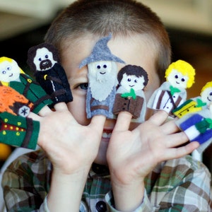 May include: A child holds up a collection of felt finger puppets, each depicting a character with distinct features. The puppets include figures with yellow, brown, and gray hair, and various outfits in green, brown, and blue. The puppets are handmade.