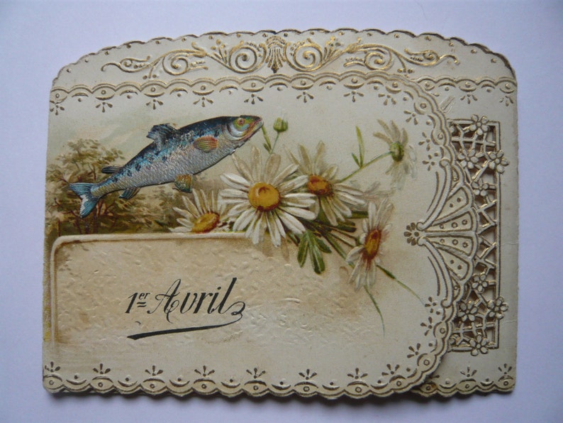 K&ouml;nnte beinhalten: Eine vintage Karte in Cremefarbe mit einem goldenen Rand und einem zarten Spitzenmuster. Die Karte zeigt ein gemaltes Bild eines Fisches, der &uuml;ber einem Strau&szlig; wei&szlig;er G&auml;nsebl&uuml;mchen schwimmt. Der Text "1er Avril" ist in schwarzer Tinte auf die Karte geschrieben.