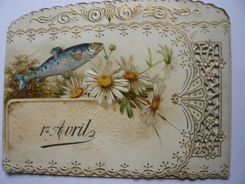 K&ouml;nnte beinhalten: Eine vintage, wei&szlig;e Karte mit goldener Umrandung und einem filigranen Spitzenmuster. Ein blauer Fisch schwimmt &uuml;ber einem Strau&szlig; wei&szlig;er G&auml;nsebl&uuml;mchen. Die Karte tr&auml;gt die Aufschrift "1er Avril" in Kursivschrift.