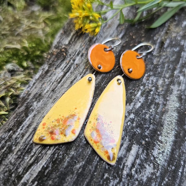 May include: A pair of handmade earrings featuring a combination of orange and yellow enamel. The earrings have a silver hook and are set on a wooden surface with yellow flowers in the background. The earrings have a unique, artistic design.