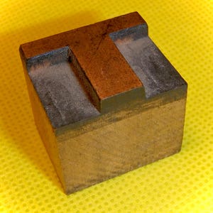 May include: A wooden letterpress block with the letter "T" carved into it. The block is brown and has a dark brown border. The letter "T" is raised and has a slightly worn surface.