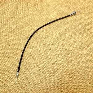 May include: A black camera shutter release cable with a silver-colored tip and a chrome-colored end. The cable is curved and lies on a tan, textured surface. This accessory is used for remote camera operation.