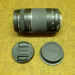 May include: A black Canon EF 75-300mm 1:4-5.6 USM camera lens with two black lens caps. The lens has a ribbed grip and measurement markings. The lens caps are round with the Canon logo.