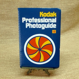 May include: A blue Kodak Professional Photoguide book with white and yellow text and a circular design in red, yellow, and white. The book is displayed on a small, decorative stand.