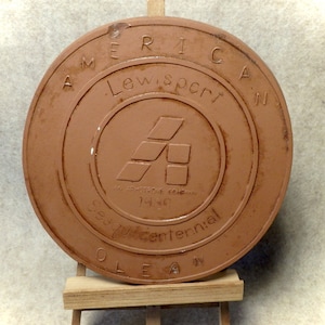 May include: A terracotta-colored round plaque with the text "America Lew,sport'n An Armstrong Company 1939 Sesquicentennial Olean" in a circle around a stylized letter "A" logo.