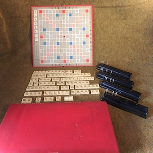 May include: A vintage Scrabble board game set with a red and blue game board, letter tiles, and a black tile rack. The game board has a star in the center and the word "SCRABBLE" printed on it.