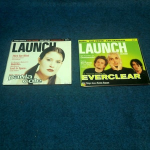 May include: Two copies of the Launch magazine, a music magazine. The cover of the left magazine features a photo of Paula Cole, a singer-songwriter. The cover of the right magazine features a photo of the band Everclear. The text on the cover of the left magazine reads "Launch" and "Paula Cole". The text on the cover of the right magazine reads "Launch" and "Everclear".