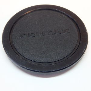 May include: A black Pentax lens cap. The cap is round with a textured surface and the brand name "PENTAX" embossed in the center. The cap is designed to protect the lens of a camera.