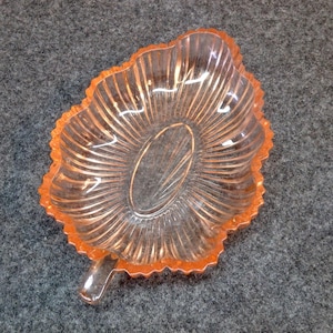 May include: A clear, leaf-shaped dish with an orange-tinted rim and a textured, ribbed design. The dish has a small stem-like handle and an oval center. The glass dish is likely used for serving small items.