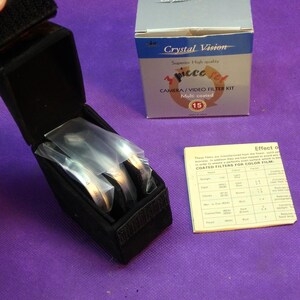 May include: A black velvet case containing three camera filters, with the text "Them 50" visible on the filters. A box labeled "Crystal Vision" and a chart detailing filter effects are also present.