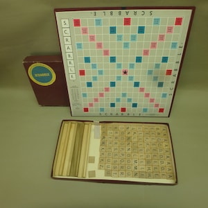 May include: A vintage Scrabble board game with a blue and red grid, a star in the center, and the word "SCRABBLE" printed on the top and bottom. The game includes a box of letter tiles, a tile rack, and a set of wooden tile holders.