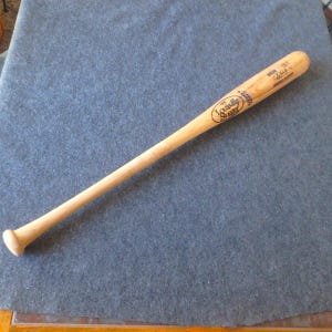 May include: A natural wood Louisville Slugger baseball bat. The bat has the Louisville Slugger logo and text printed on the barrel. The bat is resting on a blue-gray surface.