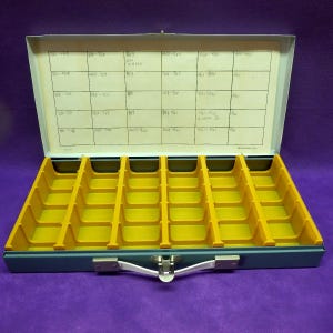 May include: A gray metal storage case with a white handle and a latch, featuring a yellow plastic insert with twenty-four individual compartments. The interior lid has a white paper grid with handwritten black ink labels in each section.