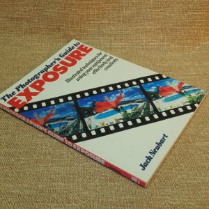 May include: A photography guide titled "The Photographer's Guide to Exposure" by Jack Neubart. The book features a white cover with red and blue text, and a film strip design with tropical imagery.