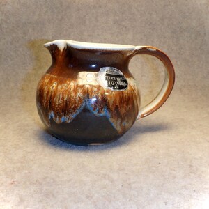 May include: A brown and white ceramic creamer with a brown handle. The creamer has a blue and white glaze and a sticker that says "Potter's Wheel Original".