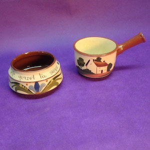 May include: Two ceramic pieces: a small sugar bowl and a handled serving dish. The sugar bowl has a brown interior and a cream exterior with blue floral accents and text. The serving dish features a house and tree design.