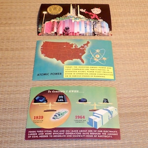 May include: Three vintage postcards depicting the history of atomic power. The first postcard shows a colorful illustration of an atomic power plant. The second postcard shows a map of the United States with a red atom symbol. The third postcard shows a scale with a large pile of coal on one side and a smaller pile of coal on the other side. The text on the postcard reads "To Generate 1 KWHR..." and shows the amount of coal needed to generate electricity in 1939 and 1964.