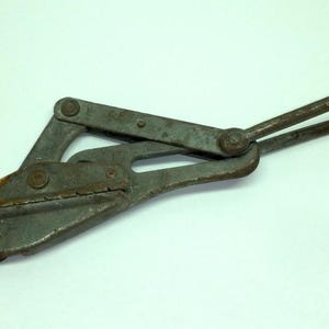 May include: A vintage, rusty metal cable grip tool. The tool has a hinged design with a loop handle and a clamping mechanism. The metal is aged with signs of wear and corrosion, suggesting it's been used.