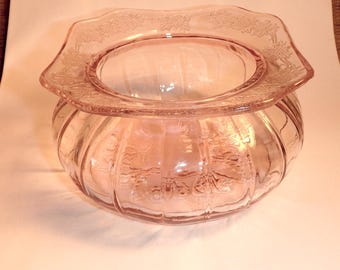 May include: A pink glass bowl with a decorative floral rim. The bowl has a round base with vertical ribbed detailing and a flared, scalloped rim. The glass has a soft, translucent appearance.