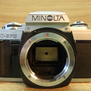 May include: A silver and black Minolta X-370 35mm film camera body. The front features the Minolta brand name on the prism housing and the model name X-370 on the left side. The lens mount is open, revealing the internal mirror mechanism.