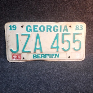 May include: Vintage Georgia license plate with white background and teal lettering. The plate reads "19 GEORGIA 83" at the top, with "JZA 455" in the center. The word "BERRIEN" is at the bottom. A small "GEORGIA 85" sticker is on the left.
