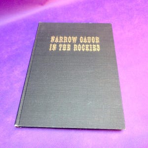 May include: A hardcover book titled "Narrow Gauge in the Rockies" with gold lettering on a dark gray cover. The book is closed and angled, set against a purple background. The cover has a textured appearance.