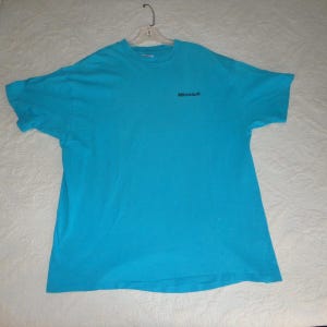 May include: A turquoise blue t-shirt with the word "Microsoft" printed in black on the upper left chest area. The t-shirt is hanging on a white hanger against a white background.